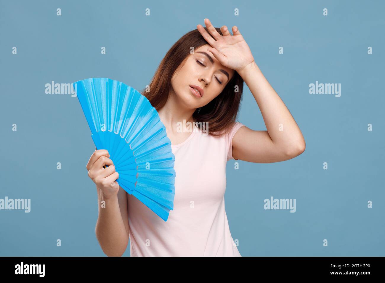 Upset millennial lady using hand fan over blue studio background, touching her forehead, closing eyes. Young pretty woman cooling herself while summer Stock Photo