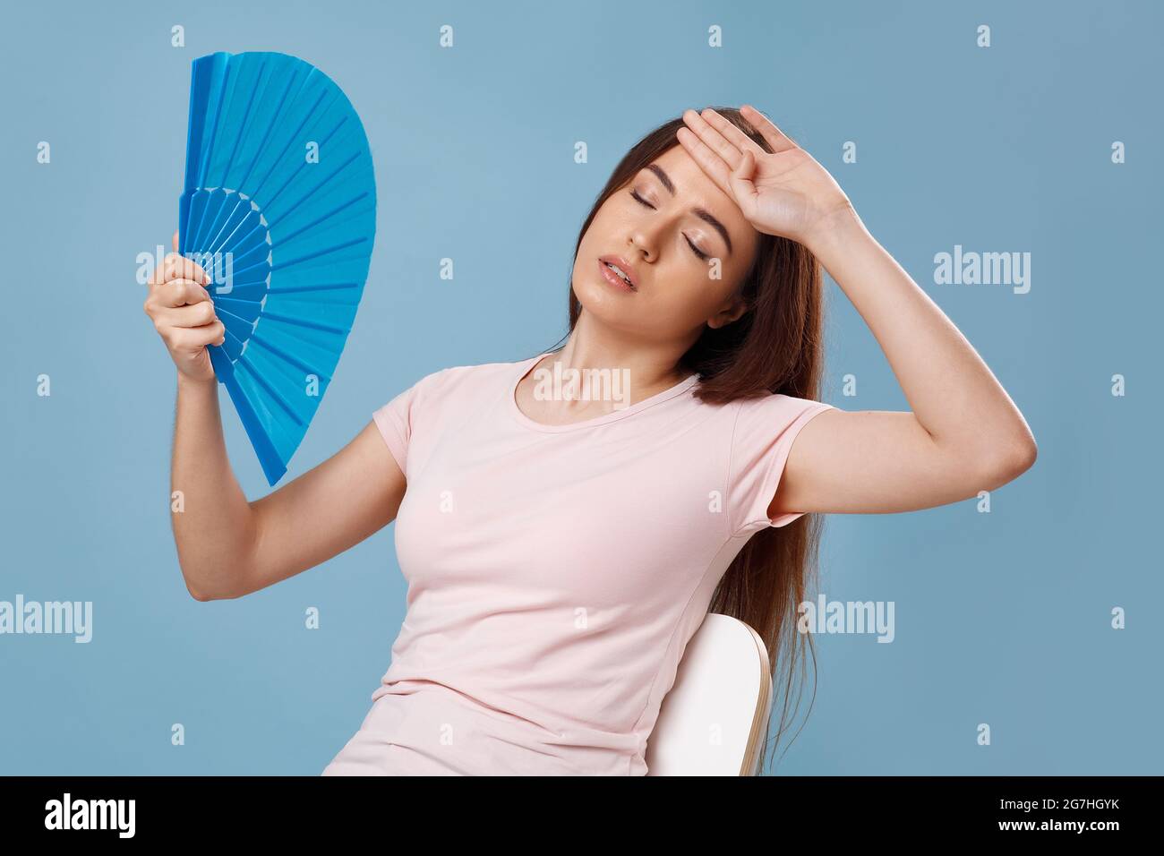 Closeup of overheated young lady in t-shirt sitting on chair with paper hand fan, pretty millennial woman suffering from heat, leaning back with close Stock Photo