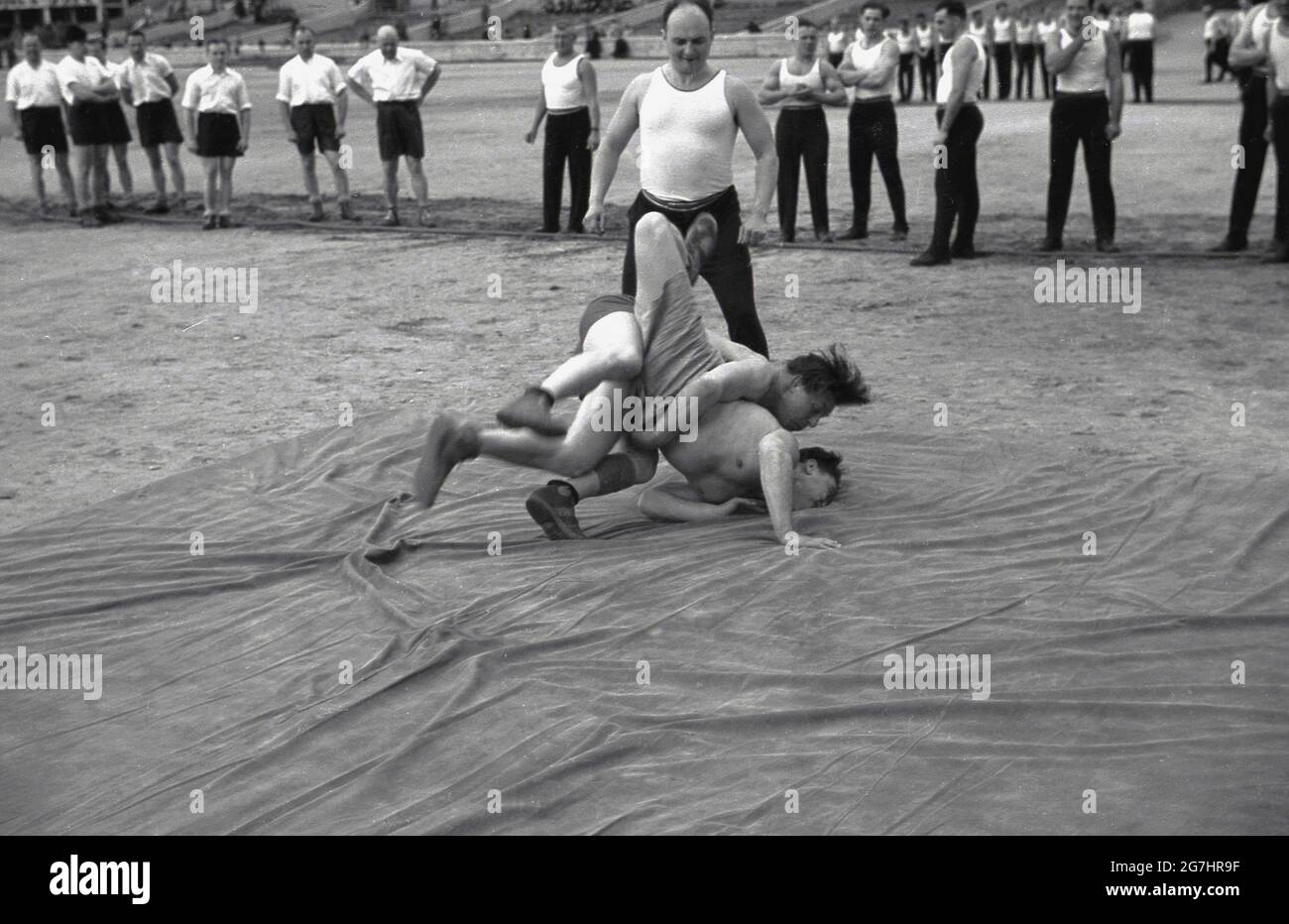 1930s, historical, inside the Strahov Stadium, Prague, Czecholsovakia, two wrestlers competiting on a large rubber mat or sheet at the Pan-Sokol Slet international festival, watched by fellow participants. Founded in 1862, the Sokol movement played an important role in the development of Czech nationalism and the early interwar years saw the Sokol flourish with high membership and large slets (mass games) Stock Photo