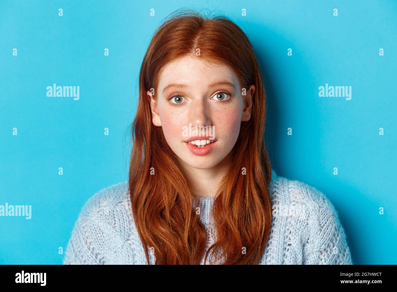 Headshot of cute redhead girl with freckles, looking hopeful and innocent at camera, standing over blue background Stock Photo