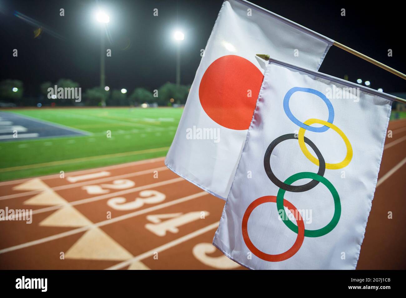 MIAMI, USA - AUGUST 15, 2019: An Olympic and Japan flag wave together under the floodlights at the starting line of a red athletics track. Stock Photo