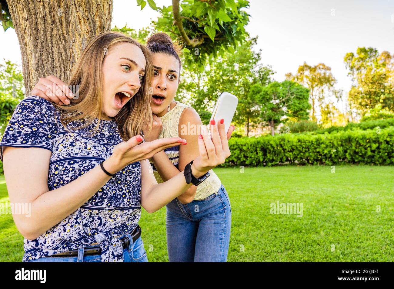 Two girls best friends look amazed at smartphone with disbelief grimaces and wide-open mouths and eyes in surprise Young women couple having fun with Stock Photo