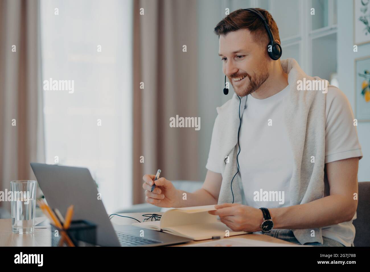 Glad male tutor gives online lessons comunicates distantly with trainee uses headphones with microphone and laptop makes necessary notes in diary Stock Photo