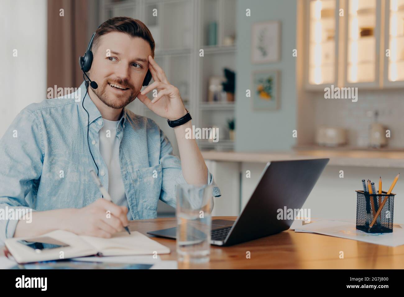 Pensive thoughtful man makes notes uses laptop computer headset writes down notes in notepad supplies customer support solves clients issues sits at Stock Photo