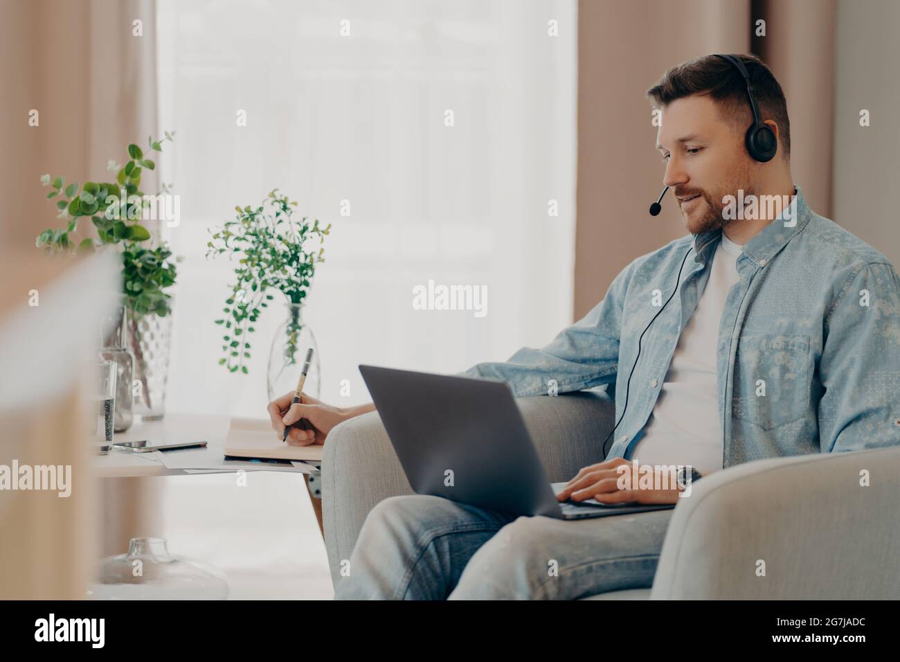 Male freelancer listens attentively tutorials how to start own business writes down information wears stereo headphones and laptop computer works Stock Photo
