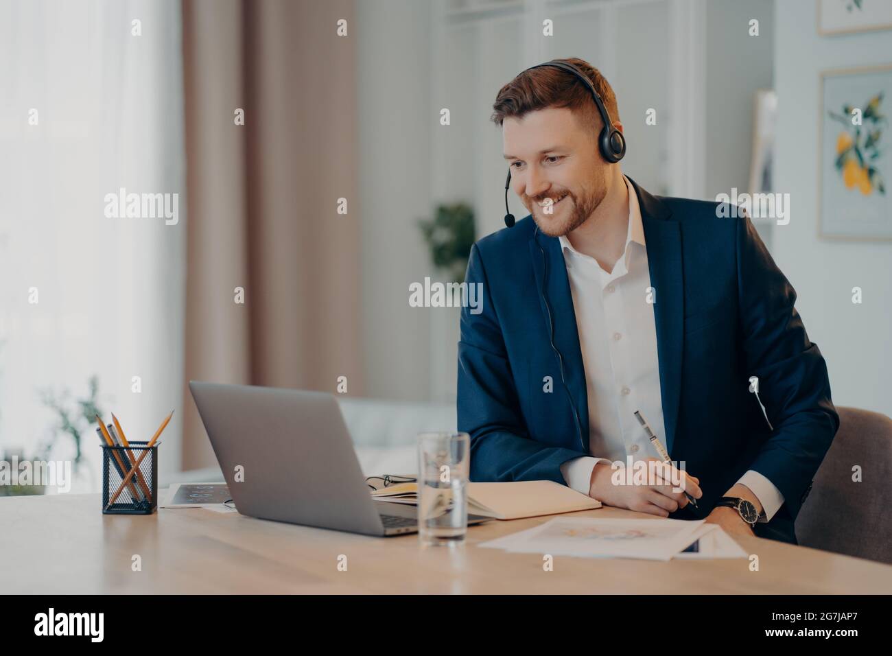 Smiling glad successful businessman practices online conference has distance meeting with colleagues wears headset focused at laptop computer wears Stock Photo