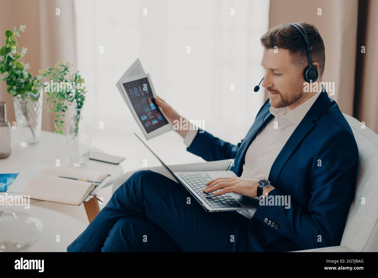 Male financial expert looks through papers with graphics dressed in formal clothes checks information on laptop computer wears headset poses against Stock Photo