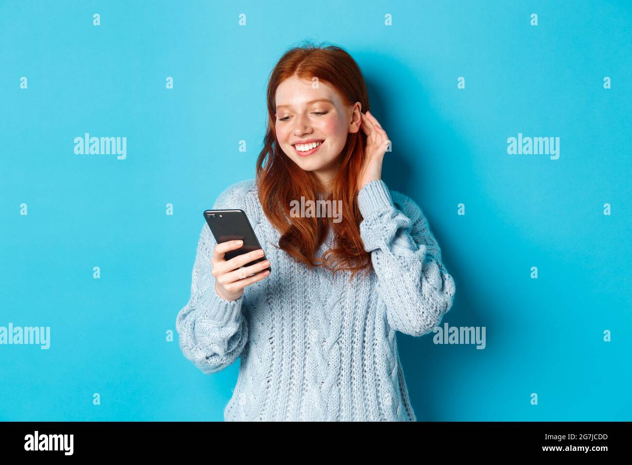 Technology. Happy redhead girl looking pleased at mobile phone, reading  compliment in message, smiling and tuck hair behind ear, standing in  sweater Stock Photo - Alamy