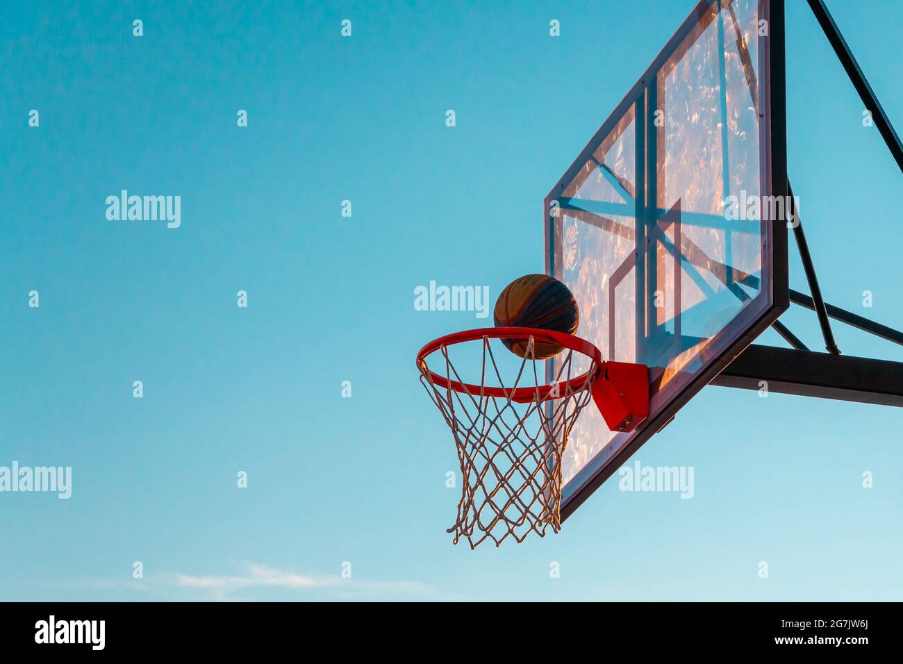 A basketball basket with a ball on a blue sky background. Transparent plastic basketball shield on the outdoor basketball court. Stock Photo