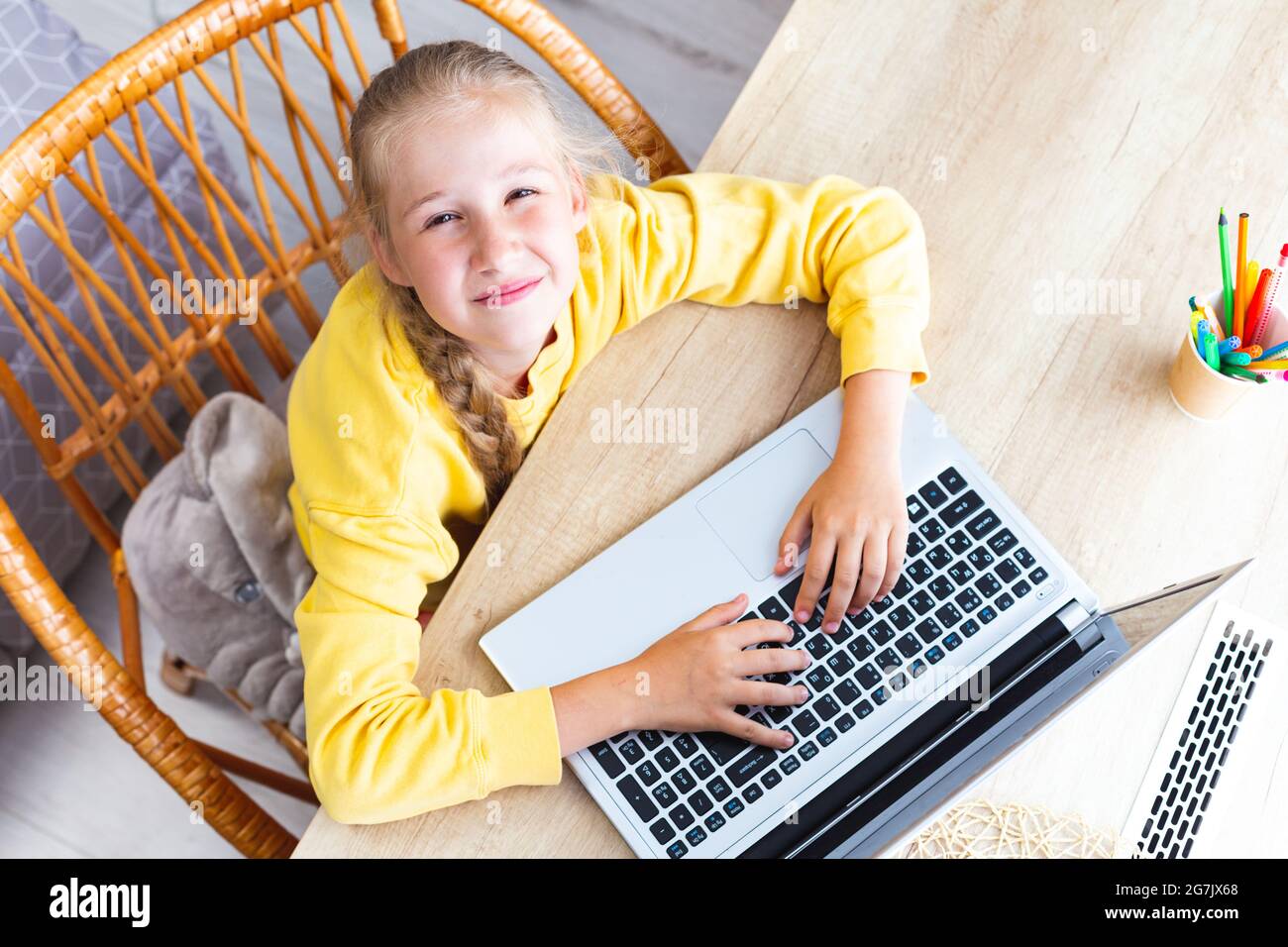 Caucasian girl 10-11 years old sits at a desk made of light wood, hands on laptop keyboard, looking at the camera squinting eyes, top view. Asthenopia Stock Photo