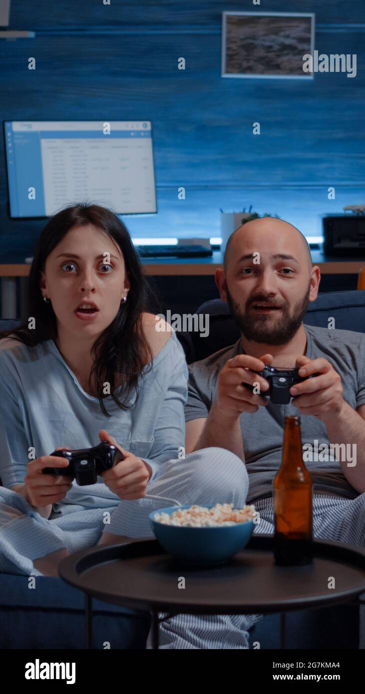 Determinated excited couple winning video games, hugged each other sitting on sofa playing, happiness scream enjoying online competition. Competitive gamers using joysticks playstation gaming Stock Photo