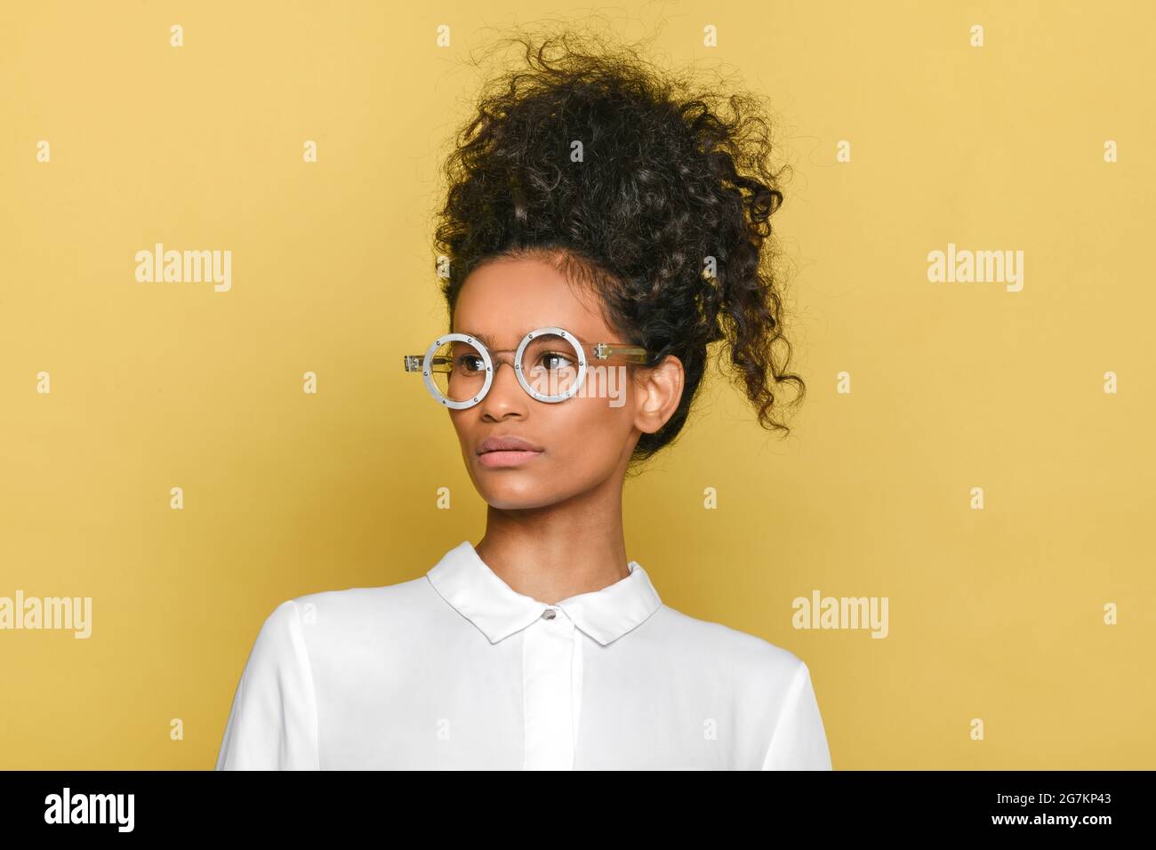 Trendy black girl in stylish retro round glasses with her long curly hair piled up on her head looking to the side over a yellow background Stock Photo