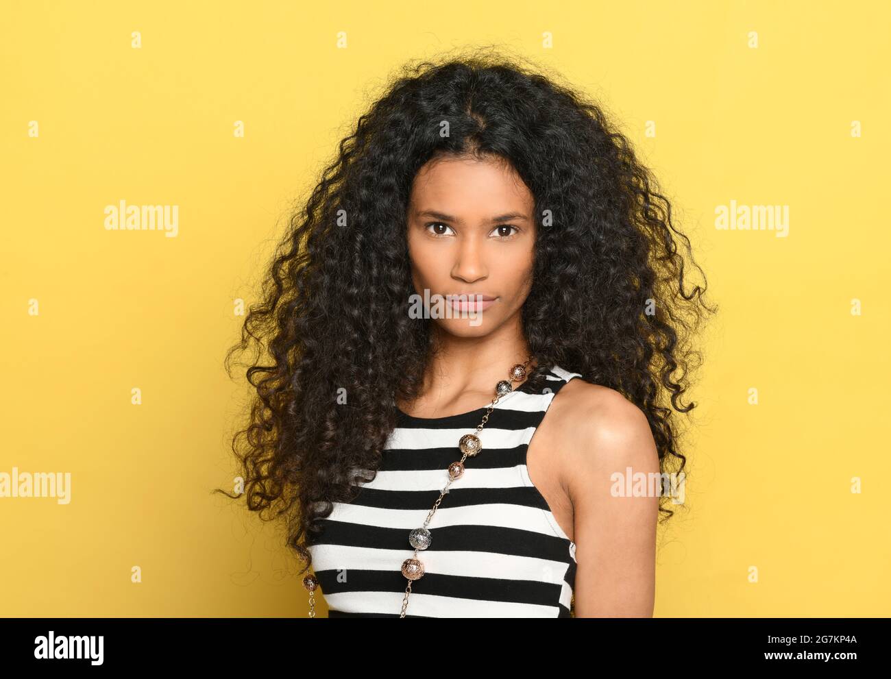 Beautiful black woman with gorgeous curly long hair posing over a yellow background looking at camera with a serious expression Stock Photo
