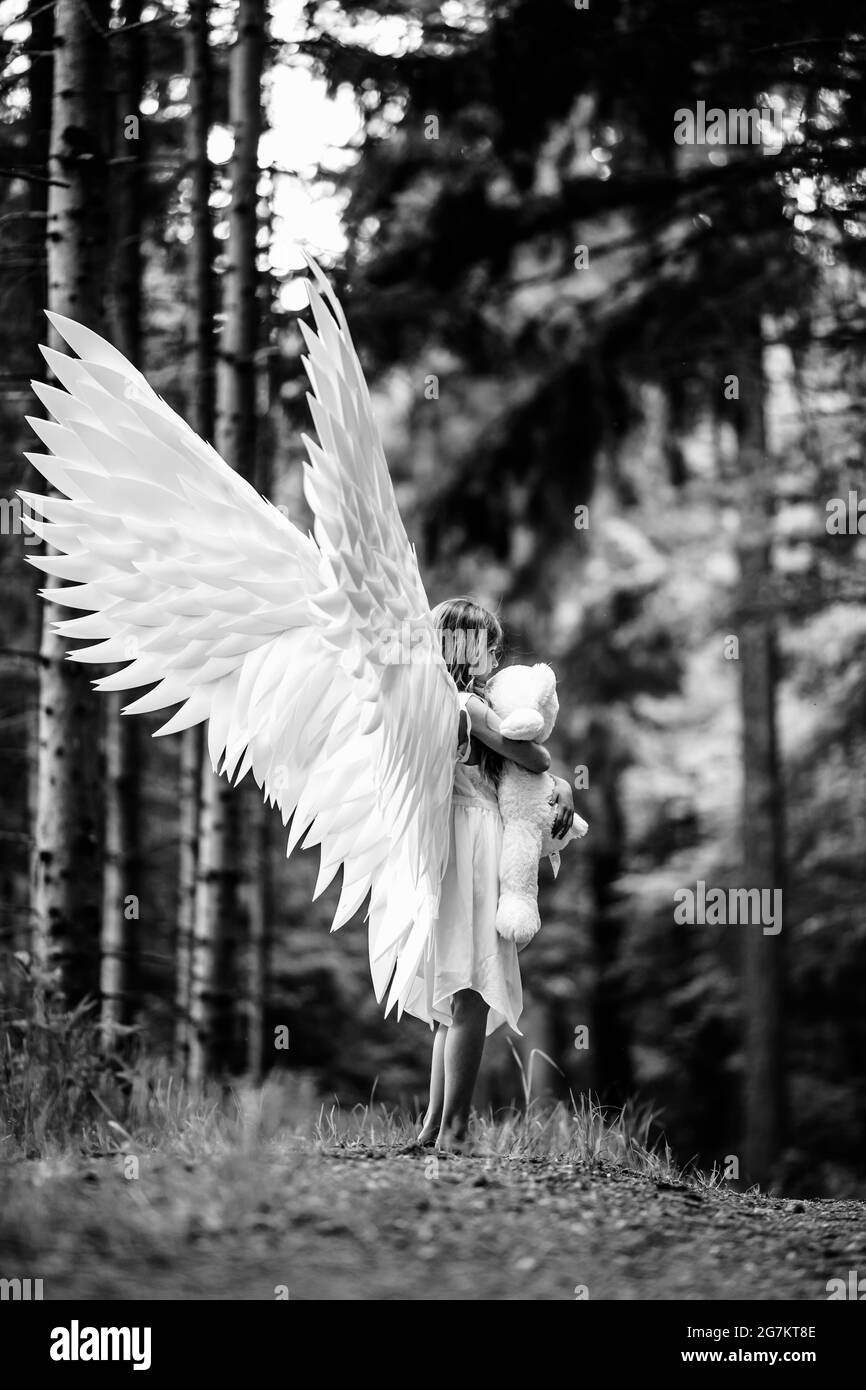 Vertical grayscale shot of a Caucasian blonde girl in angel wings costume with a teddy bear toy Stock Photo