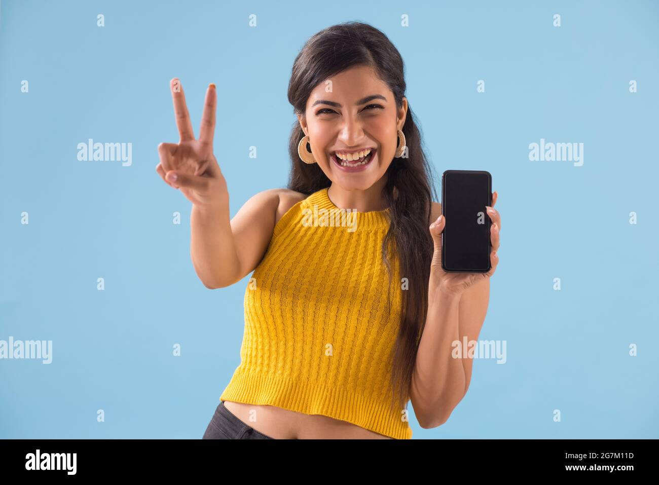 A young woman making the victory sign while showing her mobile. Stock Photo