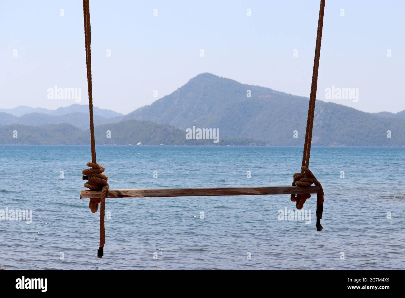 Wooden swing on a ropes against the blue sea and mountains in fog. Beach holidays on summer resort Stock Photo