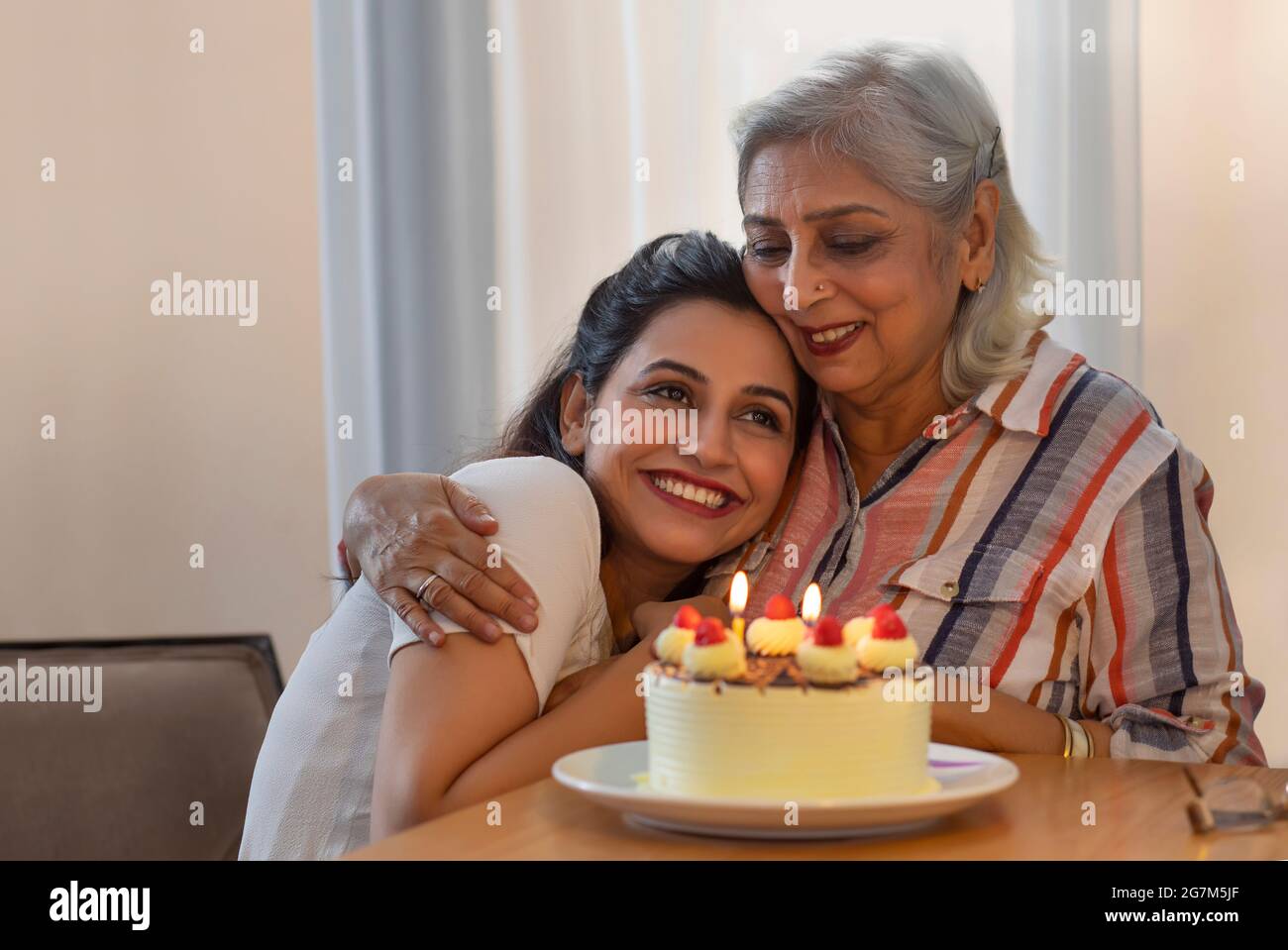 A YOUNG DAUGHTER LOVINGLY HUGGING MOTHER WITH CAKE IN FRONT OF THEM Stock Photo