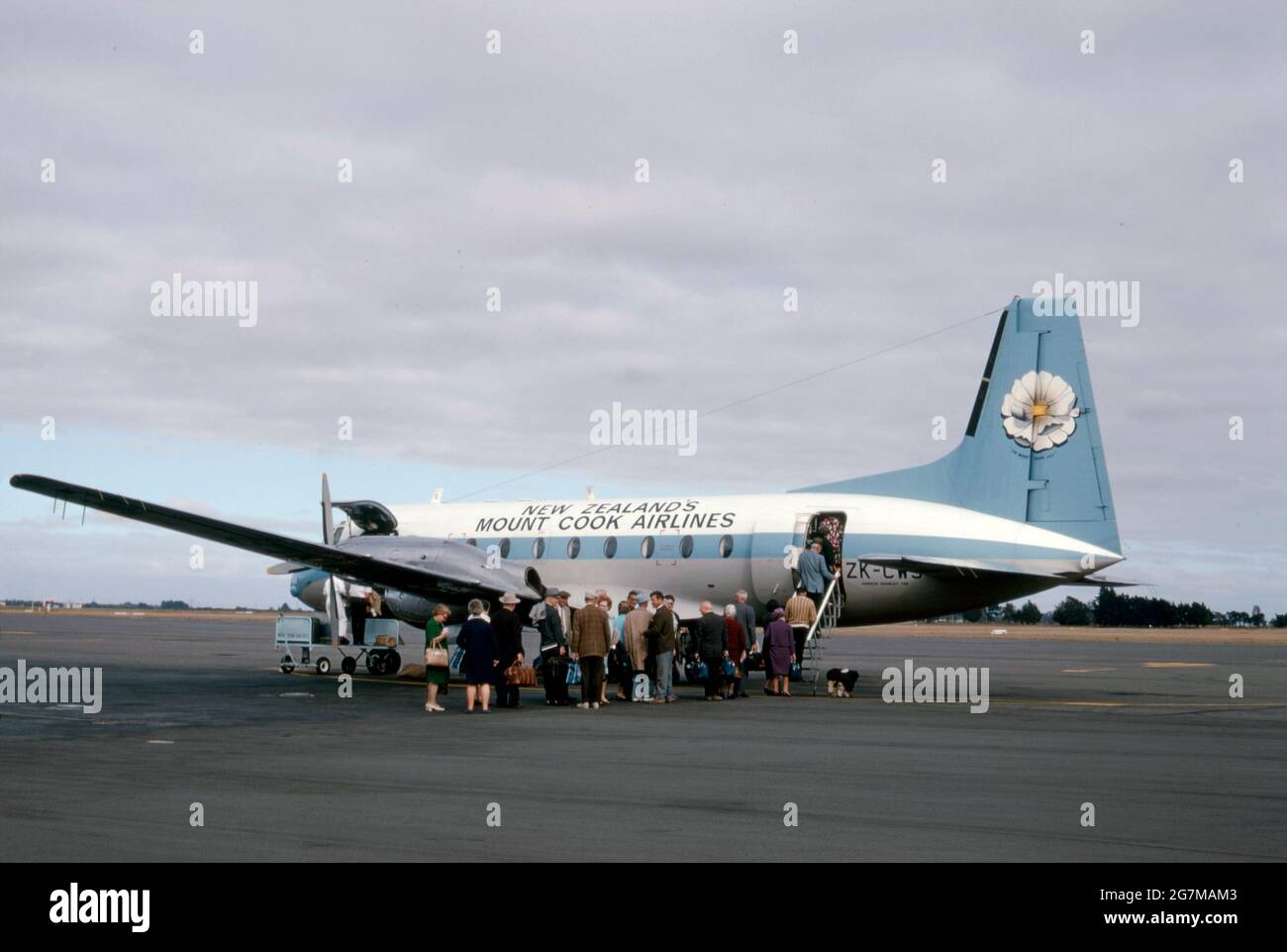 An airliner of New Zealand’s Mount Cook Airlines with passengers boarding on the tarmac at Christchurch Airport in 1969. Mount Cook Airline was a regional airline, founded in 1920, based in Christchurch, New Zealand. Formerly part of the Mount Cook Group and latterly a subsidiary of Air New Zealand, in 2019 the brand name was retired with all services flying under the Air New Zealand banner. The aircraft is a Hawker Siddeley HS 748, a medium-sized turboprop airliner. On the tail is an image of the Mount Cook lily, the largest buttercup in the world – a vintage 1960s photograph. Stock Photo