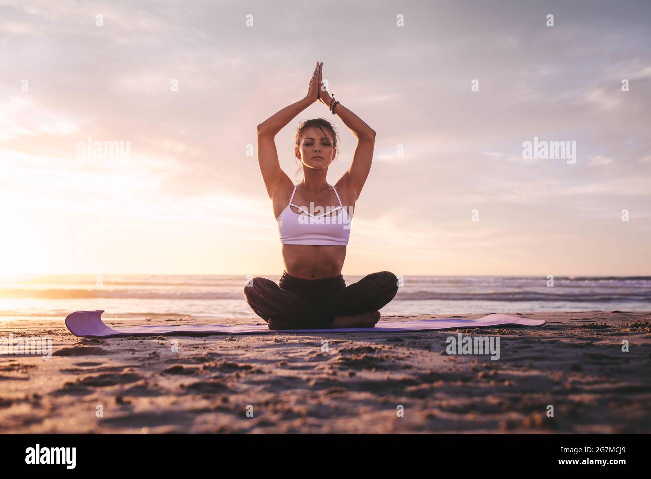 Woman in meditation on the beach. Young female sitting on the beach in lotus pose doing relaxation exercise during sunset. Stock Photo