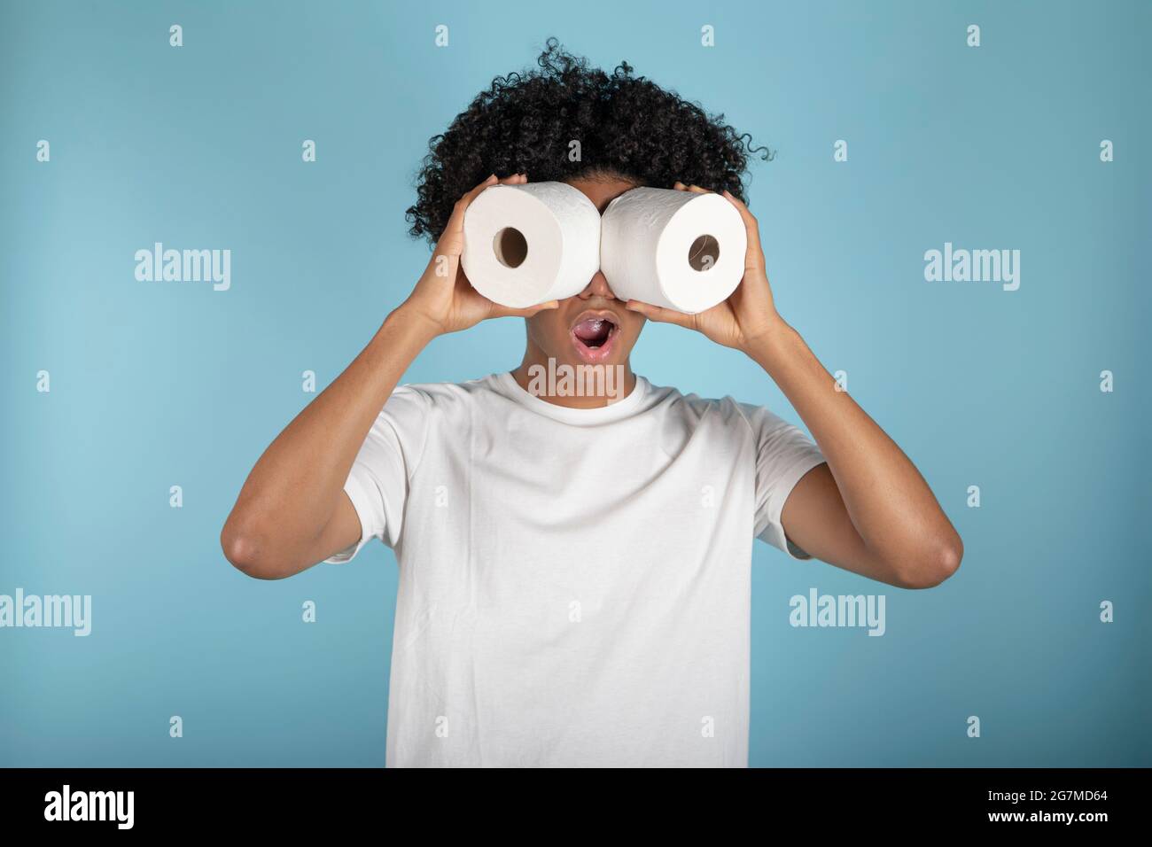 Afro-haired young man in basic white t-shirt looks astonishingly through toilet paper rolls, simulating binoculars. Isolated on blue background. Stock Photo