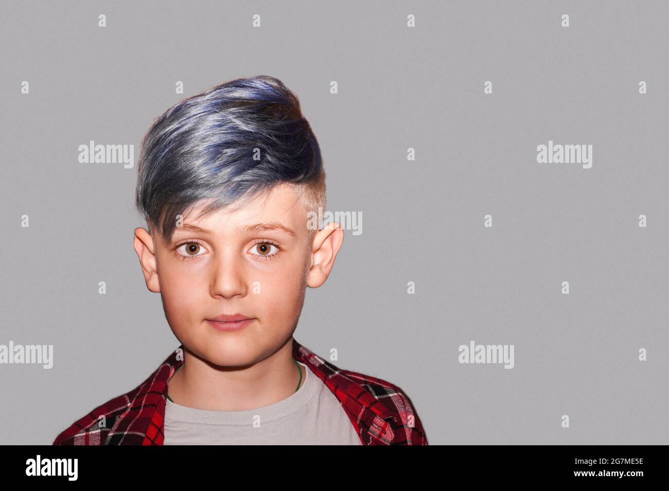Smiling happy young child with a beaming smile on gray background. Blond teen boy with Fancy trendy Hairstyle blue hair. Schoolboy standing looking at Stock Photo