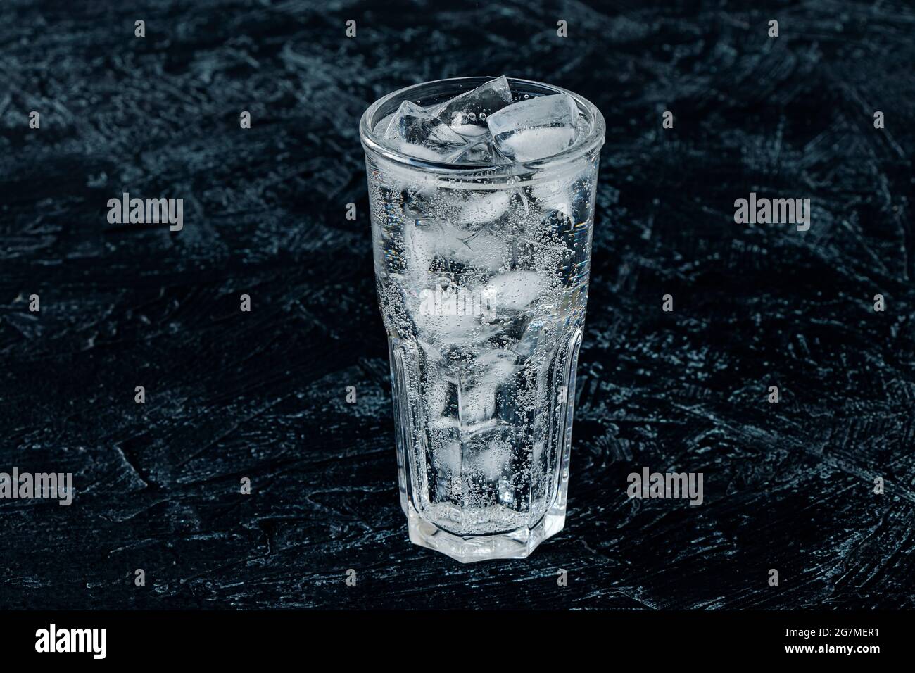 A glass with ice water and ice cubes on a black textured background. A ...