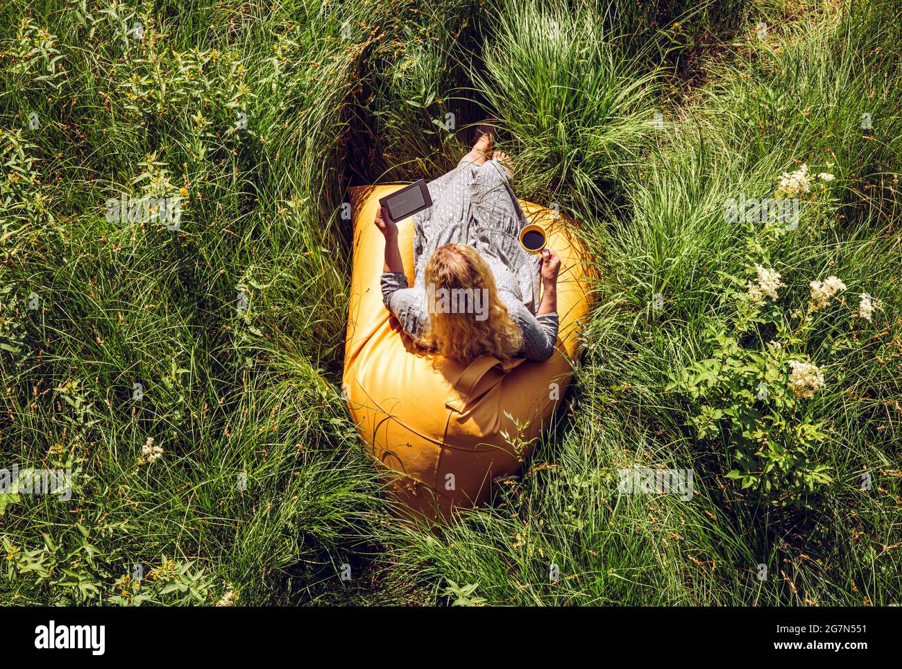 Woman person reading an ebook electronic book and sit on yellow beanbag in wild nature. Above view. Stock Photo