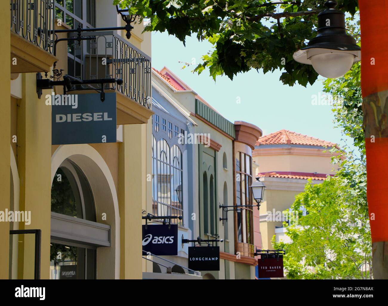 Las Rozas outlets shopping mall with signs for Diesel, Asics and Longchamp  with orange banding on tree trunks on a sunny summer afternoon Madrid Spain  Stock Photo - Alamy