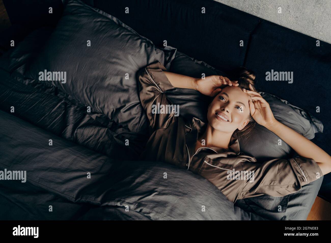 Beautiful young girl in satin comfy pajama waking up while lying in bed at home Stock Photo