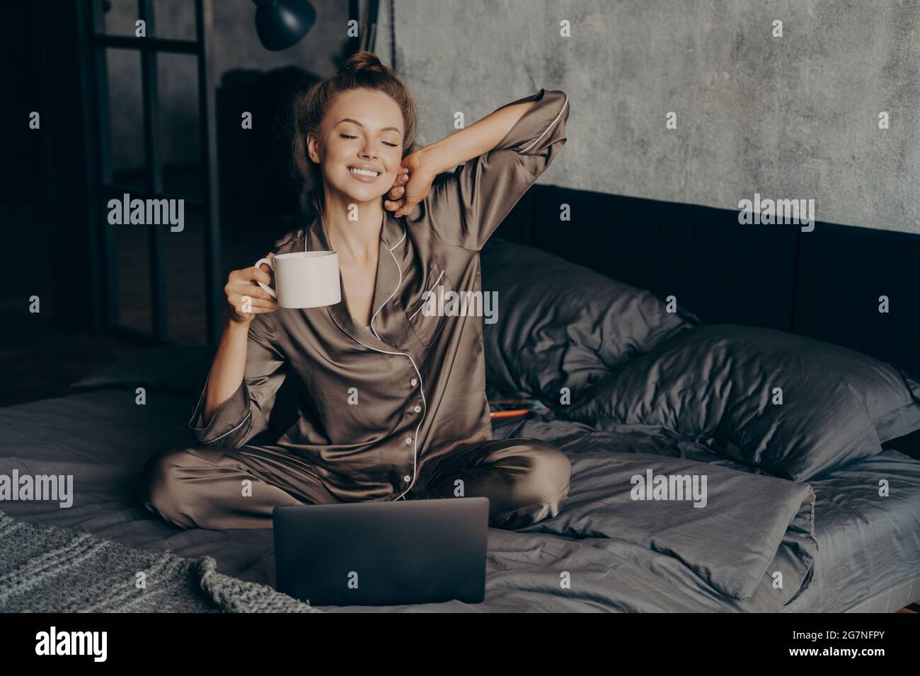 Relaxed young brunette female in satin pajama having her cup of coffee in bed and checking emails on laptop Stock Photo