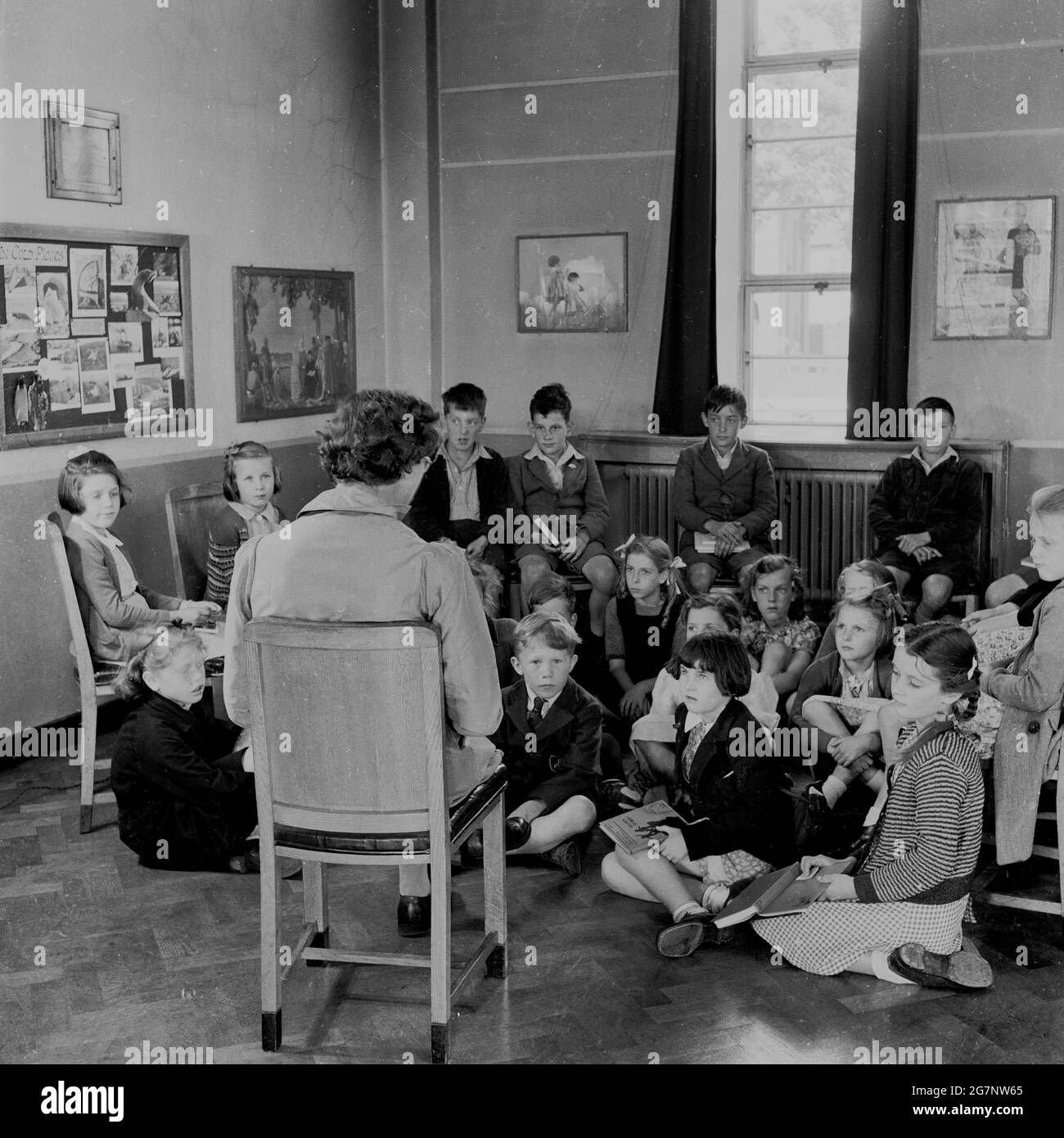 1950s, historical,  school, in the corner of a classroom, a female teacher sitting on a chair, reading a story to a group of young school school children, most of whom are sitting on the wooden parquet floor, England, UK. Stock Photo