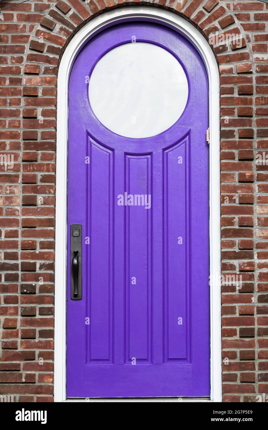 Purple, wooden, front door with a unique, circular, glass window and a rounded top, on a brick house. Stock Photo
