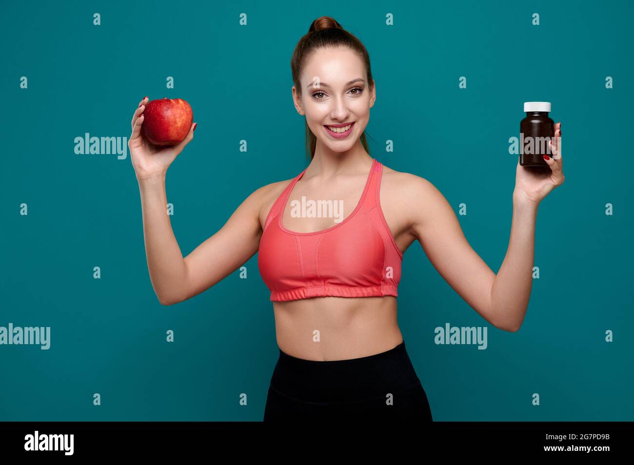 Smiling female wearing sport fit holding bottle with pills or drugs and red apple on isolate green background Stock Photo
