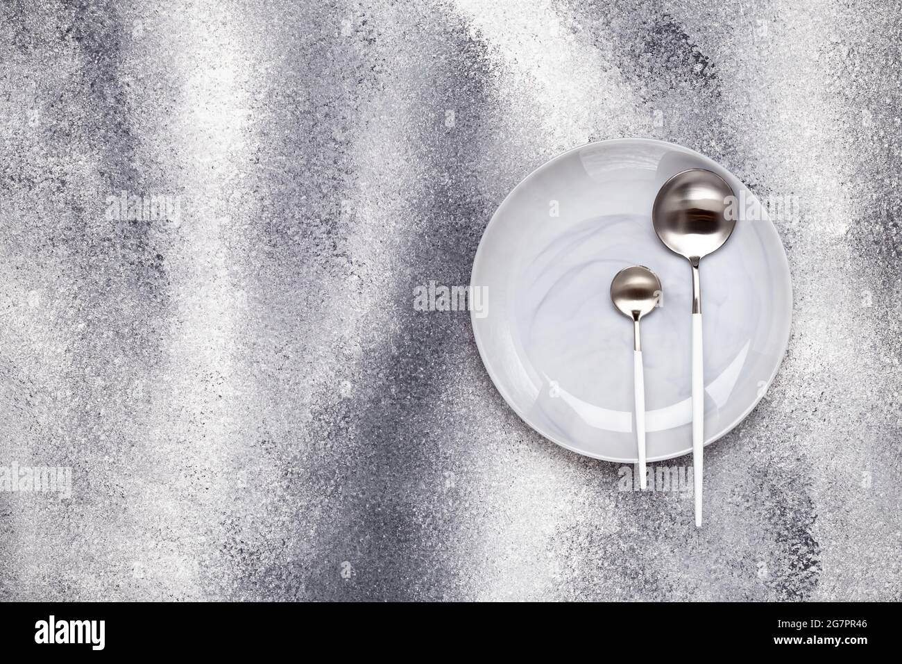 Empty clean ceramics plate and unused teaspoon with tablespoon. Crockery and cutlery on a gray grunge table, serving concept. Monochrome design menu. Stock Photo