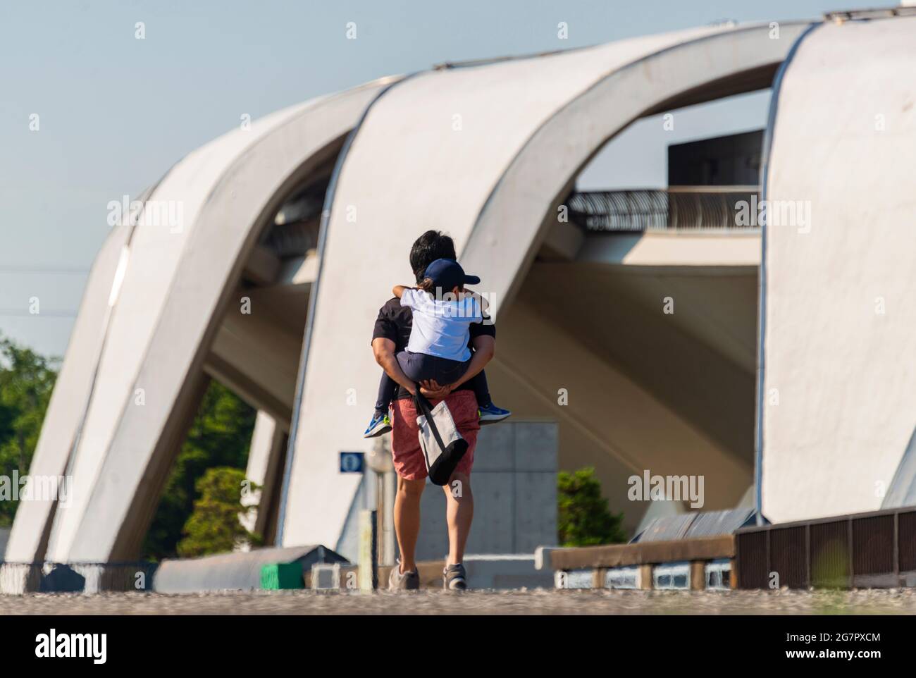 A man carries his son past the Masachika Murata-designed athletics stadium inside Komazawa Olympic Park, Tokyo on 10 June 2021. The park was built for the 1064 Olympics and remains a popular leisure venue. Robert Gilhooly photo Stock Photo