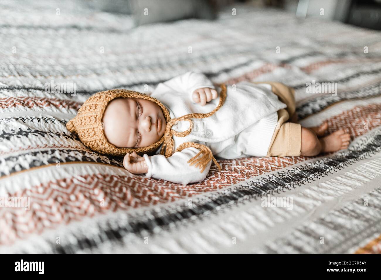 Reborn baby doll lying on a bed with a patterned dovet cover wearing a bonnet, model release not required as baby is a lifelike doll Stock Photo