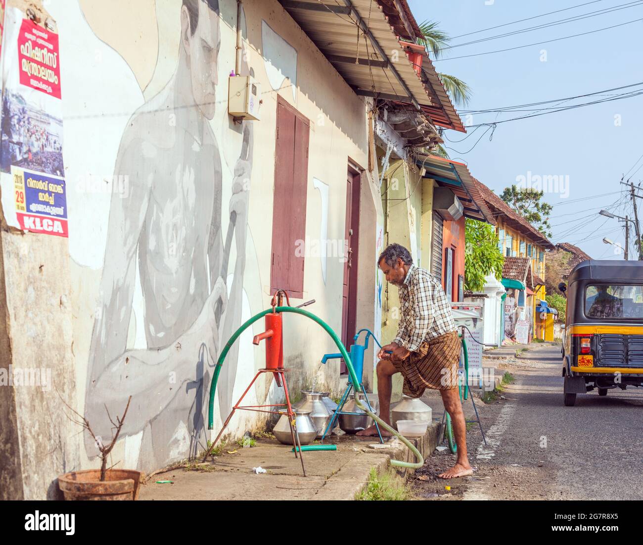 Indian male using portable water fountain to pump water into metal canisters beneath artwork on street outside home Fort Kochi (cochin), Kerala, India Stock Photo