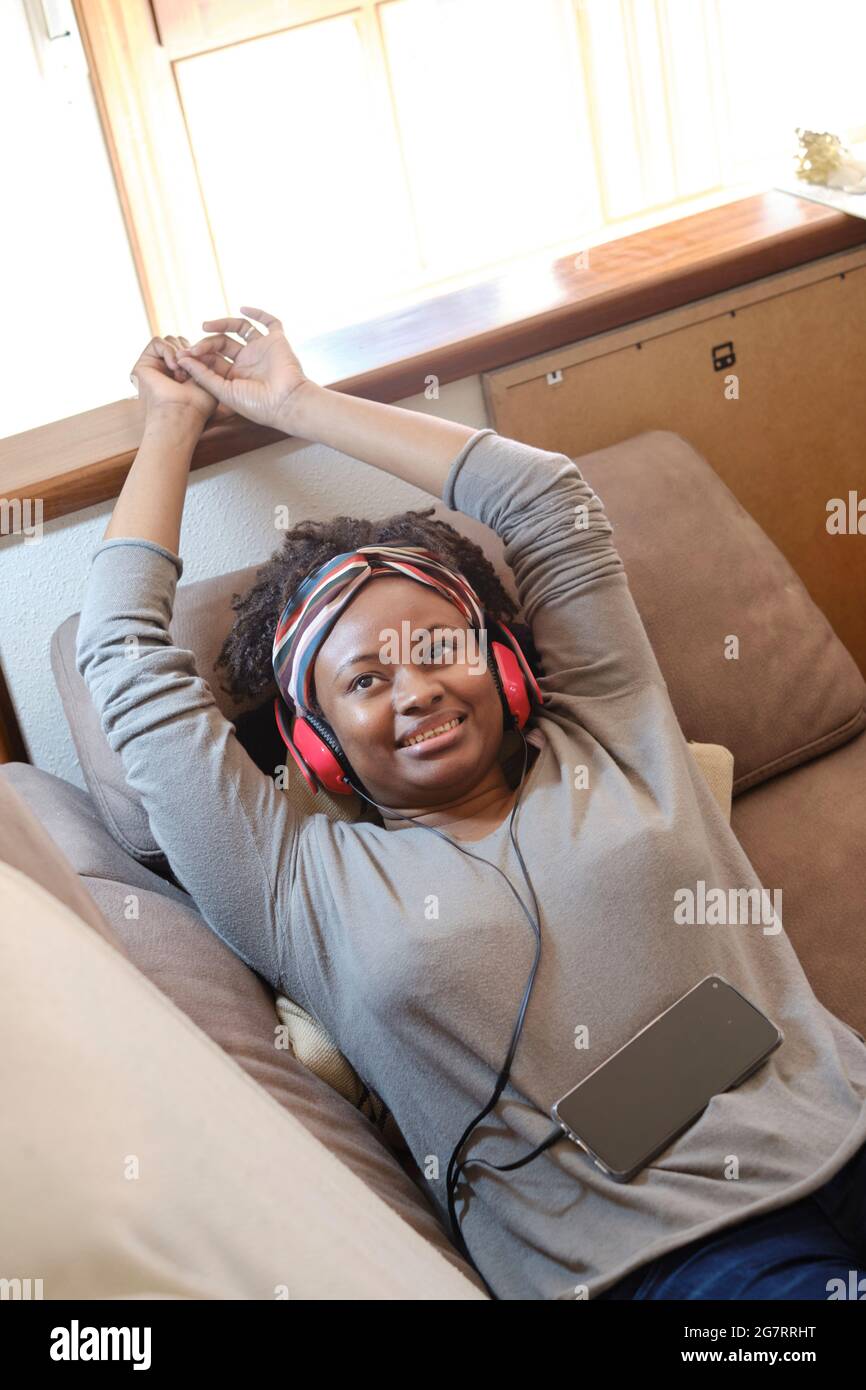 Portrait of a young black woman listening music with headphones indoor lying on a sofa.  Lifestyle concept. Stock Photo