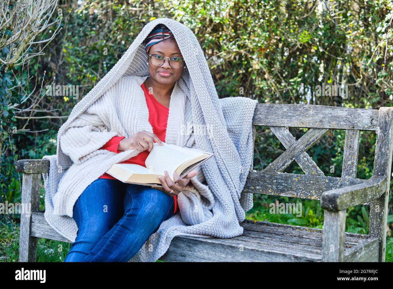Portrait of a young black woman with afro hairstyle and glasses reading a book sitting on a garden old bench. Lifestyle concept. Stock Photo