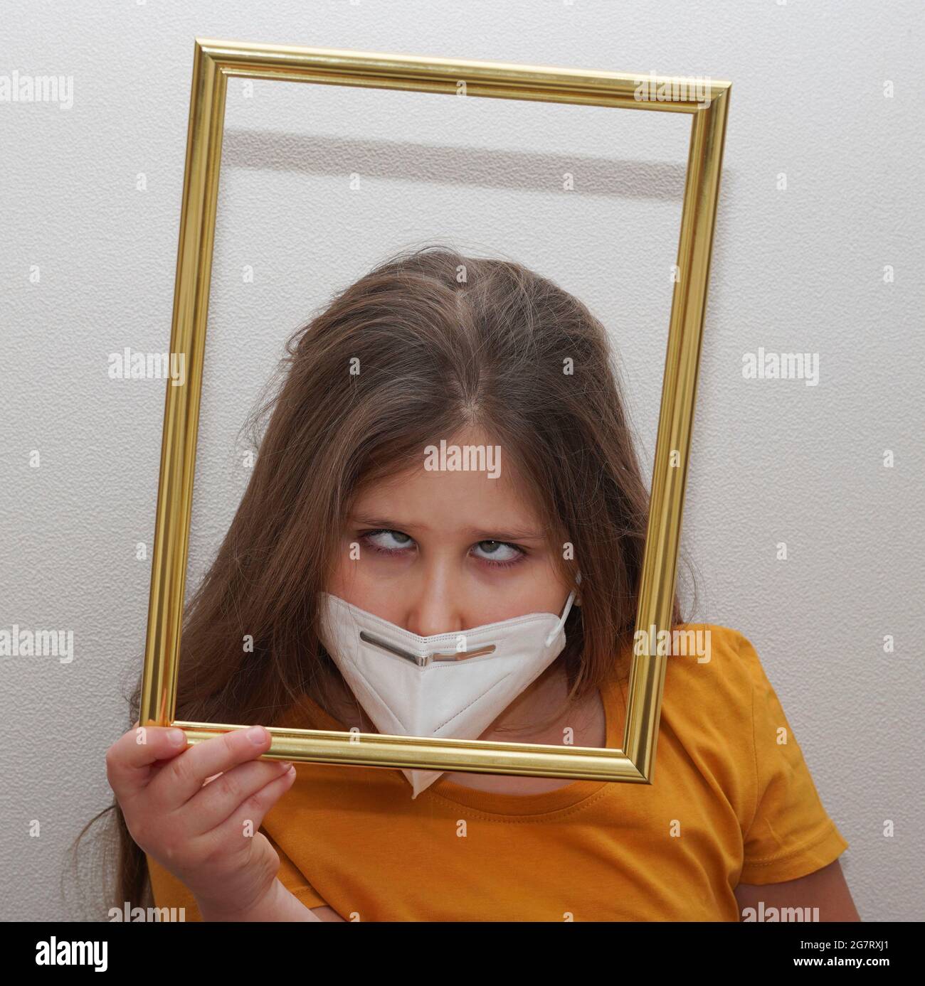 Cross-eyed girl in medical face mask and photo frame. Tired of quarantine and isolation. Protection against coronavirus and other viruses. Stock Photo