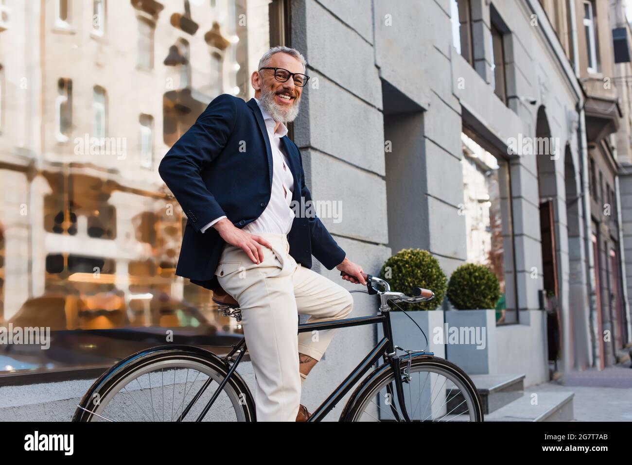 smiling bearded man in glasses posing with hand on hip and riding bicycle on urban street Stock Photo