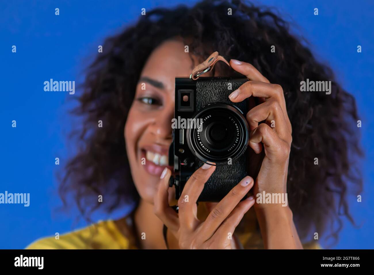 Odessa, Ukraine - June 2021. African woman takes pictures with DSLR camera - Fujifilm X100f on blue background in studio. Girl smiling as photographer Stock Photo