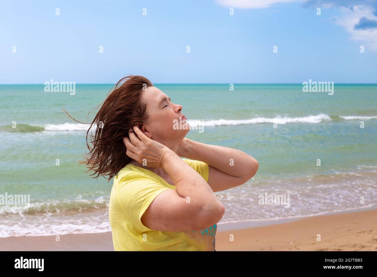 An adult elderly woman on the seashore, closed her eyes from the sea breeze. Travel and tourism in summer. Stock Photo