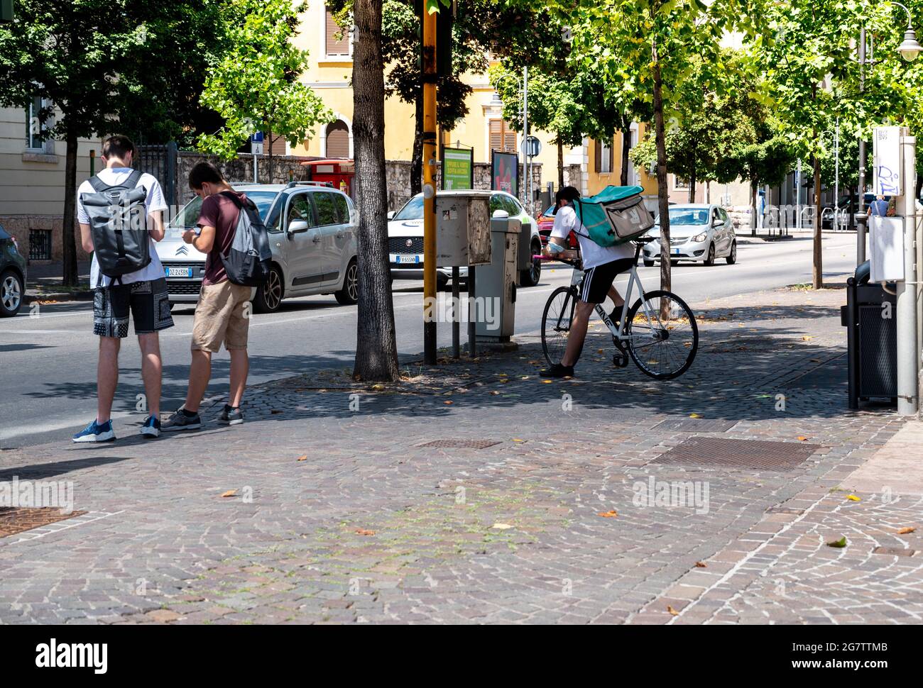 Trento, Italy, June 2021. A deliveroo worker takes a break in the shade of a tree for a break. He is consulting his smartphone. Beautiful summer day. Stock Photo