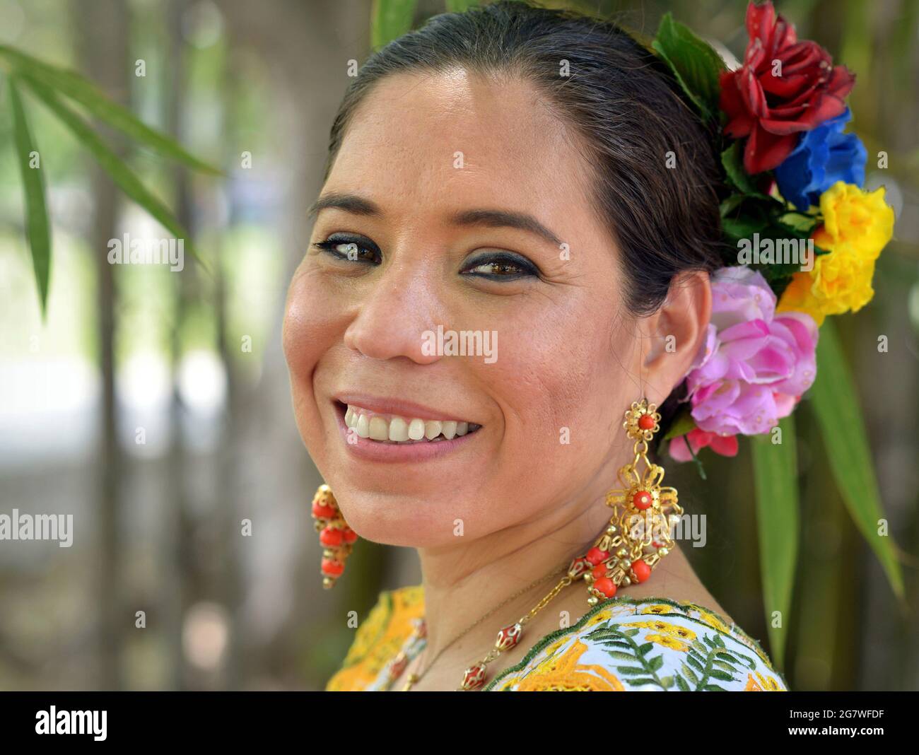 Young Mexican Yucatecan woman wears traditional Mayan folkloric dress with long earrings and colorful flowers in her hair at a park background. Stock Photo