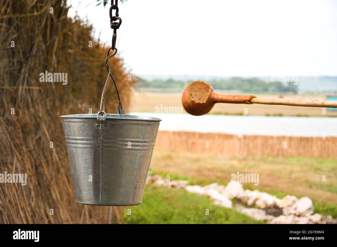 A water fountain with an old bucket and a makeshift mug made of pumpkin. Reed fence in background. Traditions and lifestyle. Stock Photo