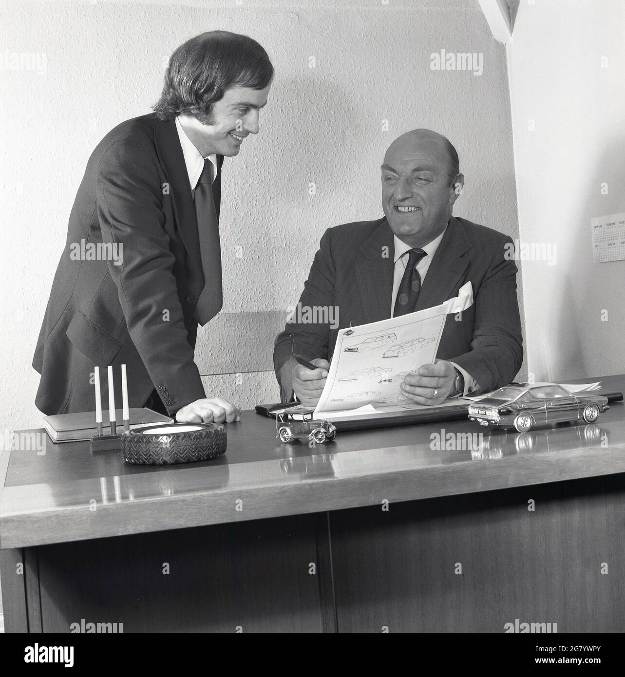 1975, historical, at a car dealership, a happy sales director sitting at his desk with his sales manager in an office, looking at a technical brochure for a new Datsun motorcar, Croydon, England, UK. They are delighted with the sales of the Japanese Datsun motorcar, the best selling imported car in Britain in the late '70s. Stylish and reliable, they were the opposite of the cars being produced by British motor maufacturer, British Leyland. Stock Photo