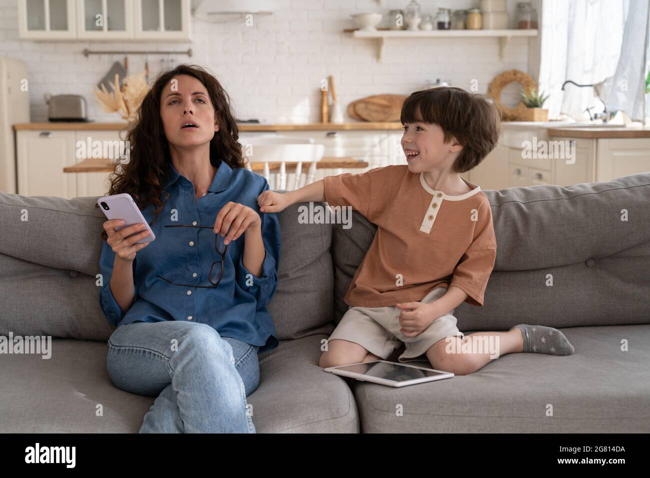 Stubborn son annoy mother at home: tired mom try to read message sit on sofa with naughty child Stock Photo