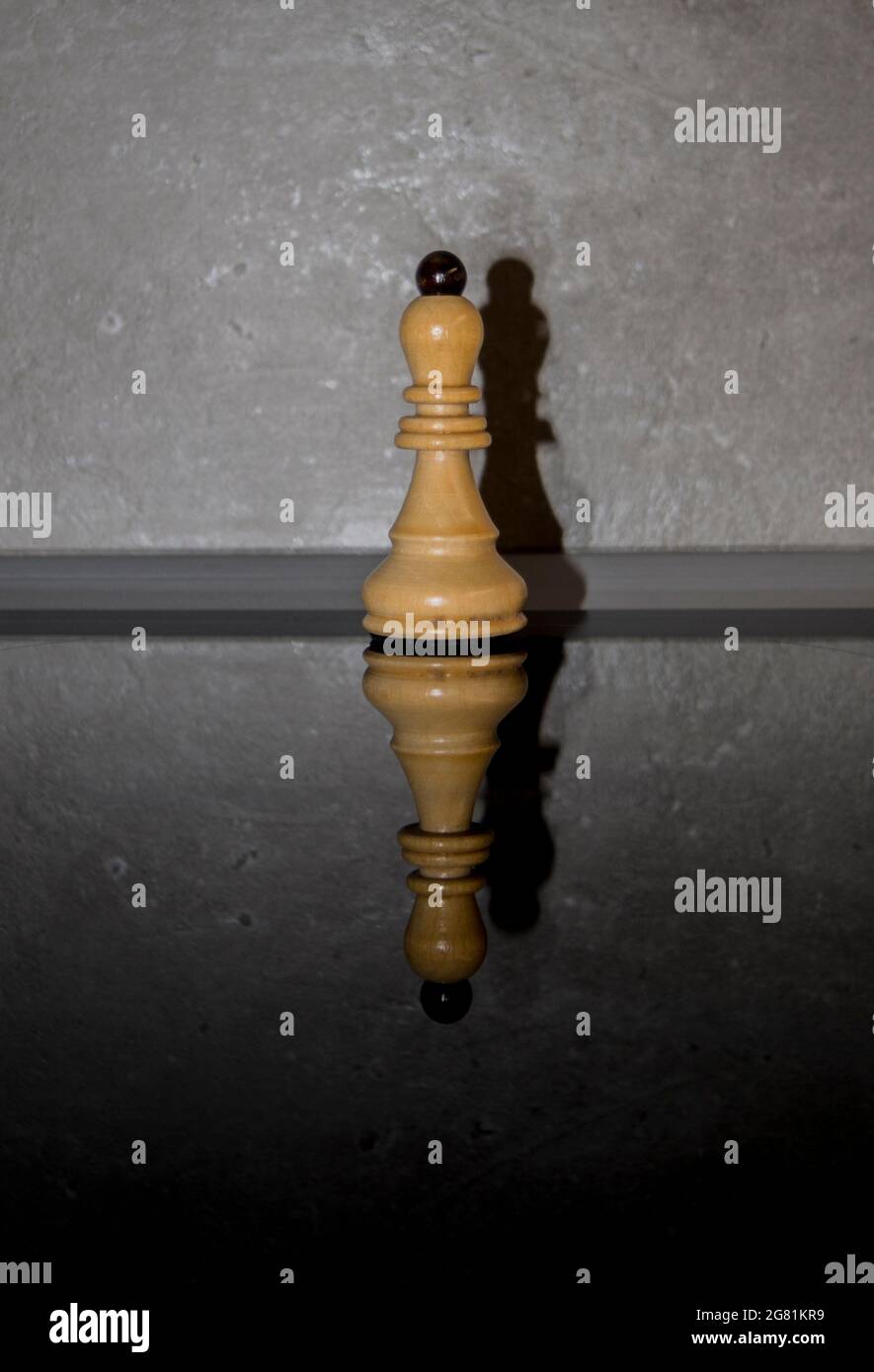Wooden white chess piece, bishop on dark mirror glass with concrete wall background. Games, sports and recreation idea. Stock Photo