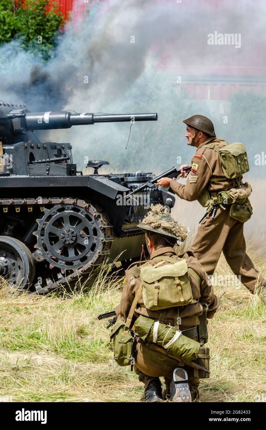 Re-enactors re-enacting a Second World War battle, with Essex regiment soldiers passing a 'burning' German Panzer tank Stock Photo